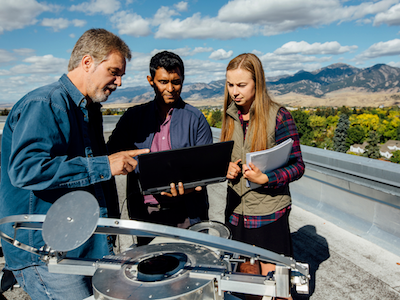 professor and two students on top of building examining computer and equipment with mountains in the background